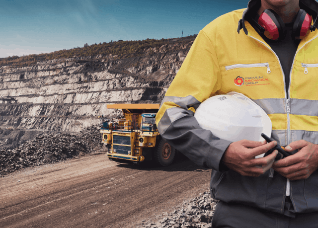 A man in a yellow jacket and hard hat stands in front of a dump truck. Relevant to mining news.